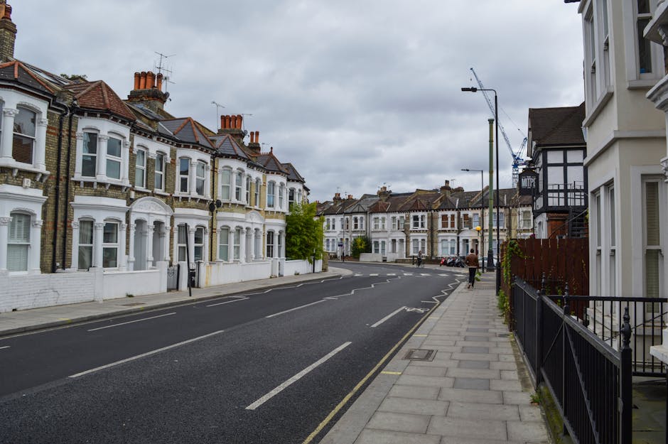 A quiet residential street in Canonbury with traditional Victorian-style terraced houses featuring white-painted facades, bay windows, and slate roofs. The pavement runs alongside the row of houses, with metal railings and small front gardens visible. The street has a few parked cars and a person walking in the distance. Overhead, an overcast sky with grey clouds provides diffused lighting. In the background, there are construction cranes indicating ongoing development nearby. The scene exemplifies typical London residential architecture, suitable for house removals and home relocation services like those offered by Islington Man and Van, who specialise in furniture transport and packing and moving within the N1 postcode area.