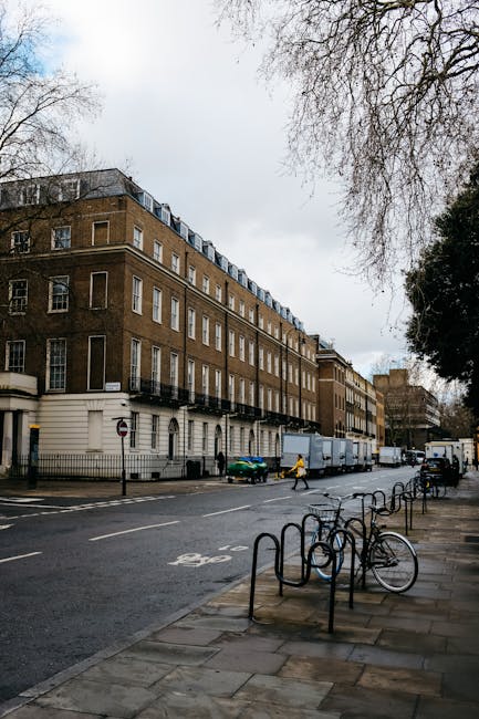 A quiet residential street in Canonbury with traditional Victorian-style terraced houses featuring white-painted facades, bay windows, and slate roofs. The pavement runs alongside the row of houses, with metal railings and small front gardens visible. The street has a few parked cars and a person walking in the distance. Overhead, an overcast sky with grey clouds provides diffused lighting. In the background, there are construction cranes indicating ongoing development nearby. The scene exemplifies typical London residential architecture, suitable for house removals and home relocation services like those offered by Islington Man and Van, who specialise in furniture transport and packing and moving within the N1 postcode area.