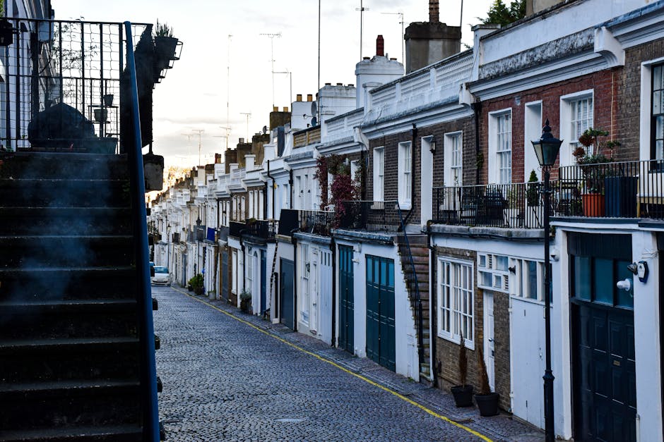 A narrow residential street in Islington lined with terraced brick houses featuring white and dark-colored doors, small front gardens with potted plants, and decorative black iron railings. The street is paved with cobblestones, and a black metal staircase on the left leads up to an upper-level property. Street lamps are mounted on the buildings, and several chimneys rise above the rooftops. The image appears to be taken during early evening or late afternoon, with soft natural light illuminating the scene. This setting exemplifies typical London home architecture, relevant to house removals and furniture transport services, such as those provided by Islington Man and Van, especially when navigating properties with narrow access for moving logistics.