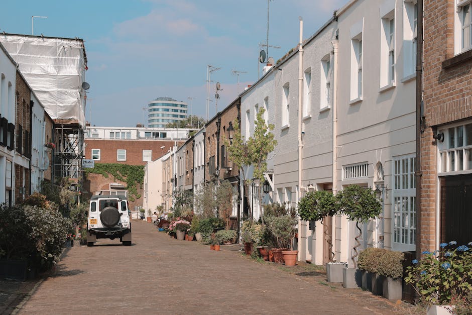 A narrow residential street in Islington with cobblestone paving, lined with closely spaced terraced houses on both sides. The houses feature a mix of brick and white painted facades, many with small front gardens filled with potted plants and shrubs. Some houses have decorative window frames and small balconies. A white van with a spare wheel mounted on the back is parked on the cobbled street, indicating the presence of a home relocation or furniture transport service by Islington Man and Van. The scene shows a loading process with some items, such as cardboard boxes and wrapping materials, possibly being prepared for moving. A few tools and equipment, like lifting straps and blankets, may be visible near the van or on the ground, supporting the logistics of packing and moving furniture through narrow access. Overhead, the sky is partly cloudy, and in the background, there are modern high-rise buildings and antennas seen above the rooftops, contrasting with the traditional terraced housing and indicating an urban setting conducive to house removals in properties with limited access.