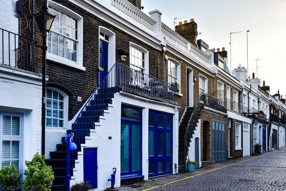 A narrow residential street in Islington lined with terraced brick houses featuring white and dark-colored doors, small front gardens with potted plants, and decorative black iron railings. The street is paved with cobblestones, and a black metal staircase on the left leads up to an upper-level property. Street lamps are mounted on the buildings, and several chimneys rise above the rooftops. The image appears to be taken during early evening or late afternoon, with soft natural light illuminating the scene. This setting exemplifies typical London home architecture, relevant to house removals and furniture transport services, such as those provided by Islington Man and Van, especially when navigating properties with narrow access for moving logistics.