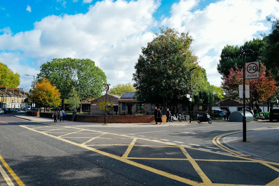 A street view outside a residential area with a small brick building surrounded by trees displaying green and autumnal foliage, under a partly cloudy sky. In the foreground, there are yellow box junct