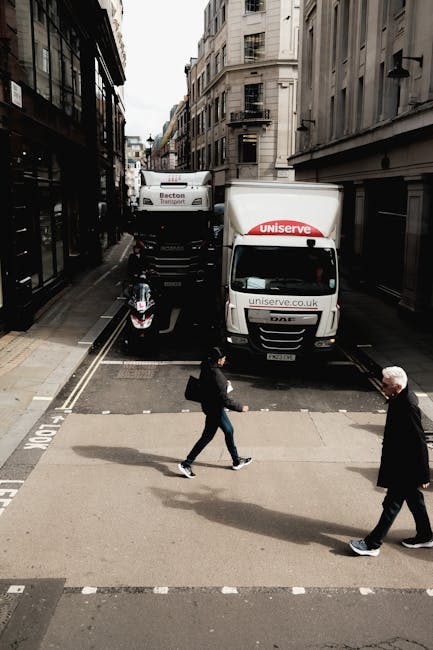 A street scene showing two individuals crossing a pedestrian zebra crossing in an urban area lined with multi-storey buildings. Behind them, a white van with 'uniserve.co.uk' branding is parked on the road, partially blocking the loading area. A larger truck labeled 'Becton Transport' is positioned further back, possibly involved in home relocation or furniture transport activities. To the left, a motorcycle is parked on the pavement, adjacent to a building with glass windows and a black framing structure. The scene is captured during daylight with overcast weather, reflecting a typical city environment suitable for house removals and moving services. The presence of the vehicles, packaging materials, and active movement illustrates the logistics involved in packing and loading during a house move, with Islington Man and Van offering professional removals in this busy urban setting.