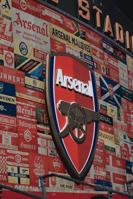 A large outdoor wall decorated with numerous patches and badges, featuring the Arsenal Football Club emblem prominently in the center. The emblem displays a red shield with a gold cannon pointing to the right, and the word 'Arsenal' written above it in bold white letters with a black outline. Surrounding the emblem are various smaller patches in red, blue, yellow, and white, representing different teams, clubs, and locations, all arranged in a collage style. The patches include text and logos related to football and Arsenal-related memorabilia. The wall appears to be part of the exterior of a building, with the collage serving as a decorative tribute to the football club. This image, as used in the context of house removals or moving services, highlights the importance of preparing and safely transporting memorabilia and collectibles during home relocation, an area where Islington Man and Van offers professional assistance.