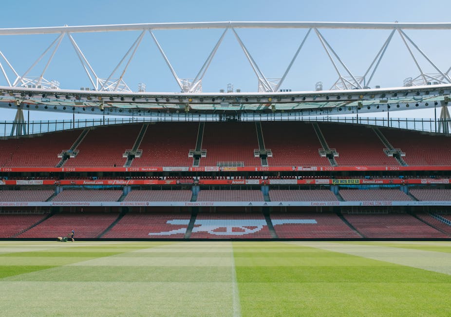 An empty football stadium featuring a well-maintained grass pitch with visible white boundary lines and a large 'A' logo in the center. The seating area comprises multiple tiers of red seats forming a continuous curve around the field, with some sections displaying sponsorship banners and advertisements, including the Arsenal Football Club logo. Above the stands, the stadium's roof is supported by a white steel framework with trusses extending across the top, allowing natural light to illuminate the playing area below. The sky is clear with a few clouds, and the overall scene is neutral and unobstructed, emphasizing the structure and layout of the sports arena. This image relates to the context of house removals and logistics, highlighting the importance of transport routes and access points for moving services, such as those provided by Islington Man and Van, especially in urban settings where parking permits for removal vans are relevant.