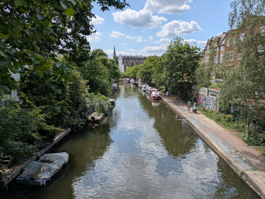 A canal scene in Islington with calm water reflecting the partly cloudy sky, bordered by a paved walkway on the right side where pedestrians are walking. Trees with lush green foliage line both sides of the canal, extending towards the background where residential buildings with traditional brick facades are visible. Several boats, some covered with protective tarps, are moored along the canal's edge. Parked cars are present on the right side of the walkway, alongside graffiti-covered walls, indicating urban surroundings. The setting captures a peaceful, residential area often associated with house removals and furniture transport logistics, with the canal providing a scenic backdrop for home relocation activities supported by local companies like Islington Man and Van.