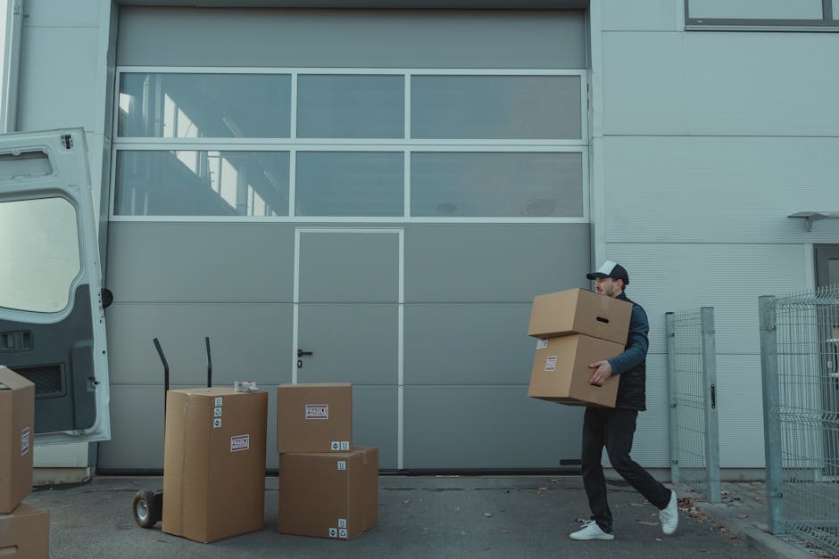 A man wearing a dark jacket, black trousers, white sneakers, and a cap is carrying a large cardboard box during a home relocation process outside a modern building with a grey exterior and large glass windows. Several other cardboard boxes of different sizes, some with labels and barcodes, are placed on the pavement nearby, suggesting packing and moving preparations. An open van with its rear door visible is parked close by, indicating the loading or unloading phase of furniture transport. A hand truck or dolly is positioned against the building wall, ready to assist in heavy items' movement. The setting appears to be an organised moving site, with the man and company, Islington Man and Van, engaged in efficient packing and logistics as part of a house removal service. The environment is well-lit, with natural daylight illuminating the scene and minimal exterior vegetation.