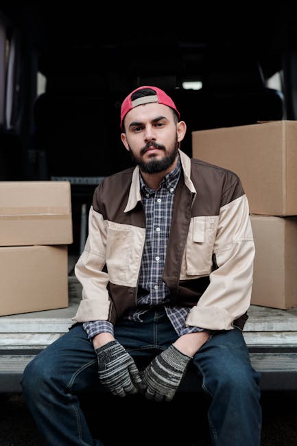 A young man with a beard, wearing a red baseball cap backwards, a brown and beige jacket over a checkered shirt, and grey gloves, sitting on the edge of a loading area at the back of a van. Behind him are cardboard moving boxes stacked around, some on the ground and some inside the vehicle. The boxes vary in size and are sealed with packing tape. The van's interior is dark with a visible ceiling and seats, indicating the vehicle is prepared for home relocation or furniture transport. The scene is set outdoors, on a paved surface, with natural lighting highlighting the loading process involved in packing and moving household items. The presence of boxes and the loading environment subtly supports the context of a professional removal service, such as Islington Man and Van, assisting with house removals or relocation logistics.