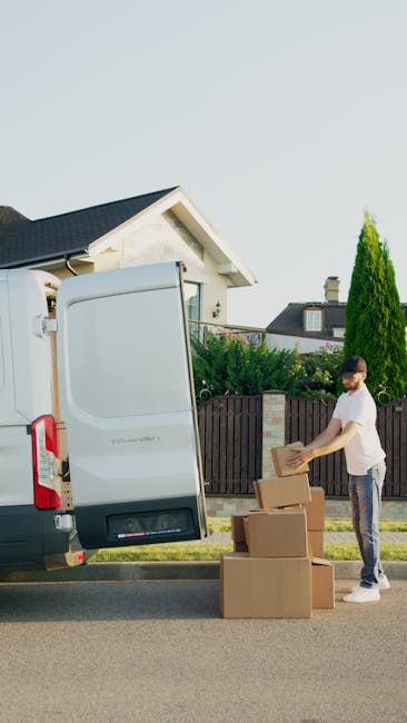 A young man with a beard, wearing a red baseball cap backwards, a brown and beige jacket over a checkered shirt, and grey gloves, sitting on the edge of a loading area at the back of a van. Behind him are cardboard moving boxes stacked around, some on the ground and some inside the vehicle. The boxes vary in size and are sealed with packing tape. The van's interior is dark with a visible ceiling and seats, indicating the vehicle is prepared for home relocation or furniture transport. The scene is set outdoors, on a paved surface, with natural lighting highlighting the loading process involved in packing and moving household items. The presence of boxes and the loading environment subtly supports the context of a professional removal service, such as Islington Man and Van, assisting with house removals or relocation logistics.
